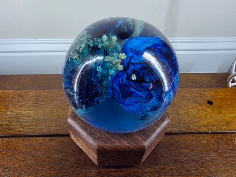 Clear resin sphere with a preserved blue rose inside, resting on a walnut base on a wooden table