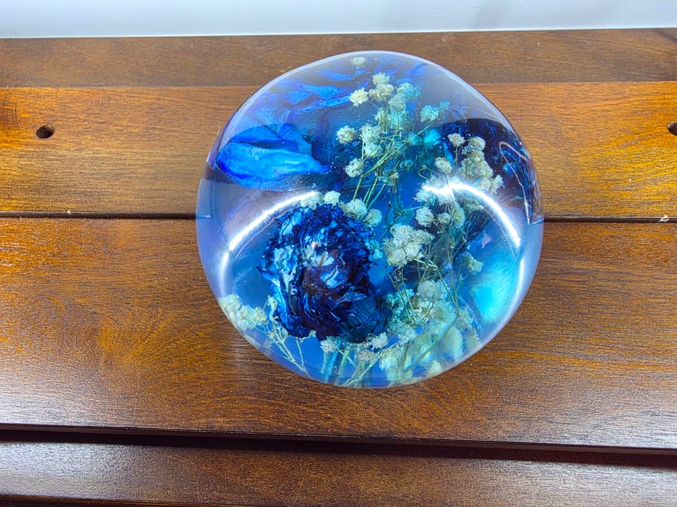 Top view of a clear resin sphere with a blue rose and small white flowers inside, resting on a wood table