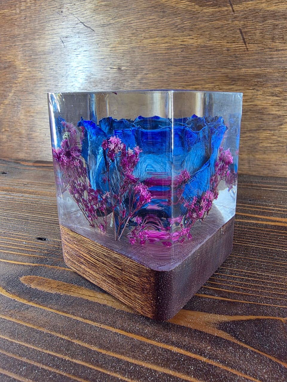 Clear resin cube holding a blue rose and magenta dried flowers, standing on a dark wood base on a plank table