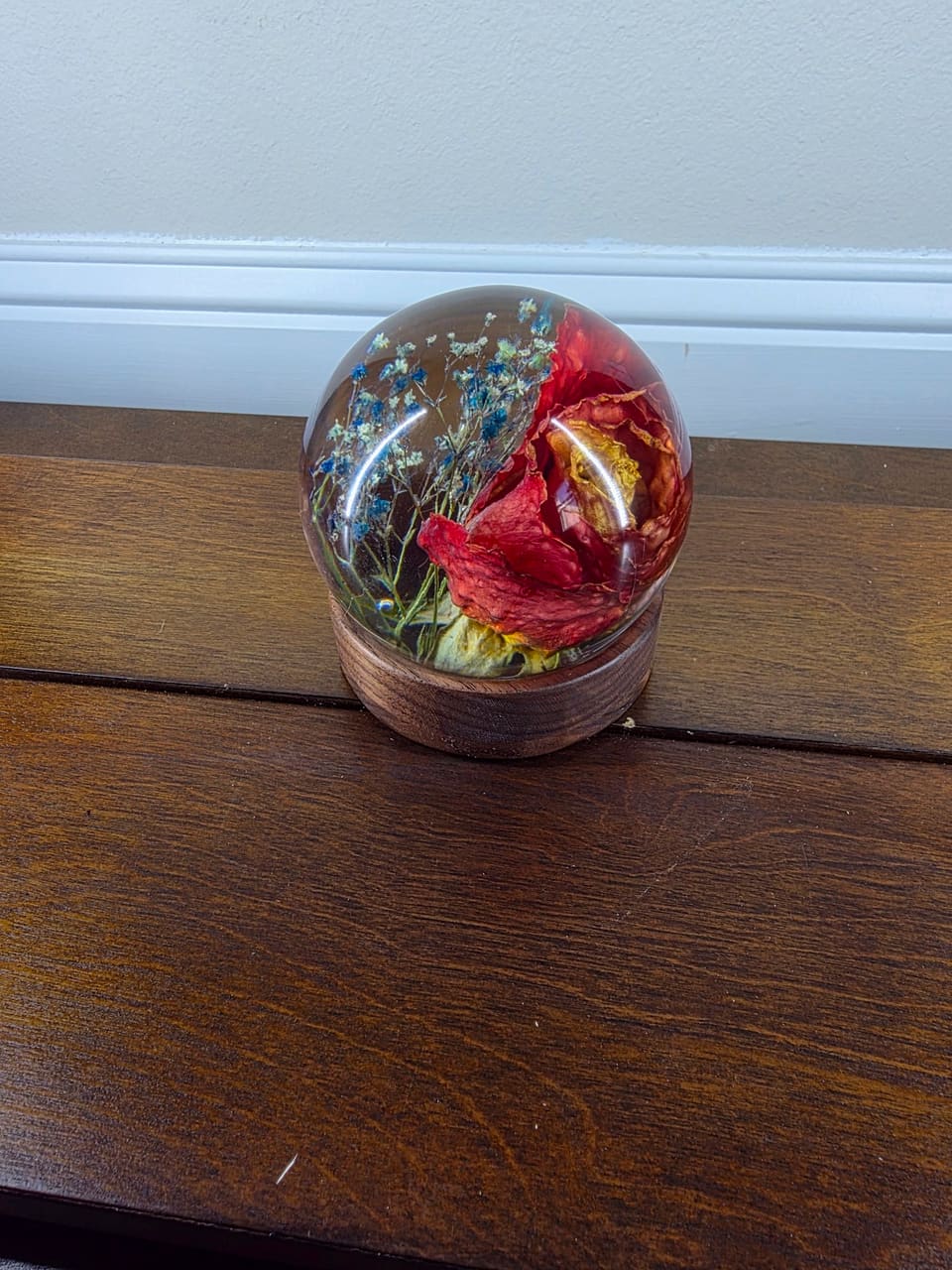 Clear resin sphere with a dried red rose and small blue flowers inside, on a round dark wood base on a wood shelf