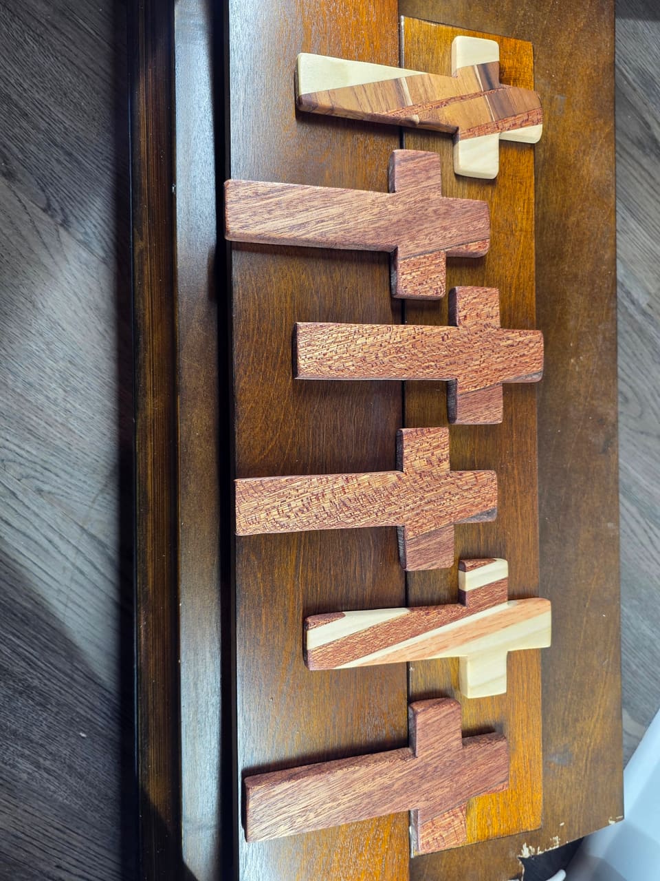 A column of small wooden crosses in two tones of wood laid on a dark wood surface