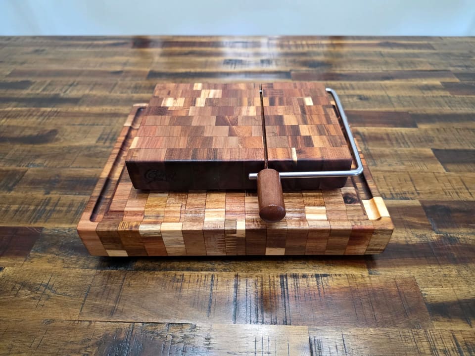 End-grain cheese slicer board with a wire and wood handle, sitting on a larger end-grain board